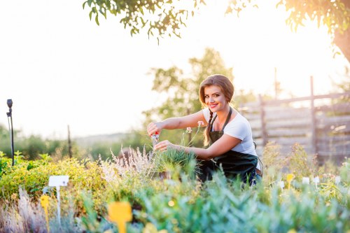 A woman working in a well-maintained garden with a variety of plants, shrubs, and flowering herbs, positioned in the foreground with lush green foliage. The garden features a mix of dense grass, flower beds with colorful blooms, and neatly pruned hedges. In the background, there are wooden fencing and mature trees providing shade, with warm sunlight filtering through the leaves, creating a bright and inviting outdoor environment. The surface includes a combination of natural soil, well-kept grass, and occasional paving areas. The woman is smiling and appears engaged in gardening activity, possibly watering or tending to the plants. The scene reflects a tidy, landscaped outdoor space that may be serviced by local gardening providers such as Garden Maintenance Wood Green, aligned with recommendations for garden upkeep in the Wood Green area, N22 postcode, highlighting a neat and vibrant garden setting suitable for routine maintenance or seasonal planting tasks.